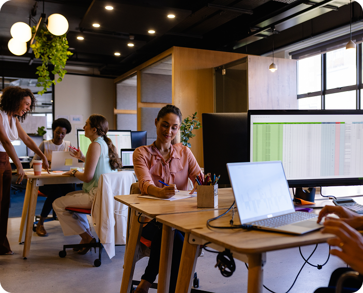 Office setting with people working at desks on computers, including a woman writing notes and spreadsheets visible on screens